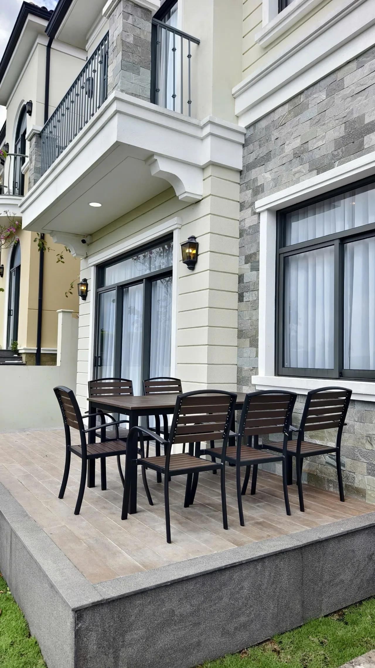 Ảnh thực tế hiên lát sàn với bộ bàn ăn sáu ghế ngoài trời, cửa kính phòng khách phía sau và bãi cỏ phía trước, căn nhà D.HOUSE bàn giao tại The Tropicana, NovaWorld Hồ Tràm