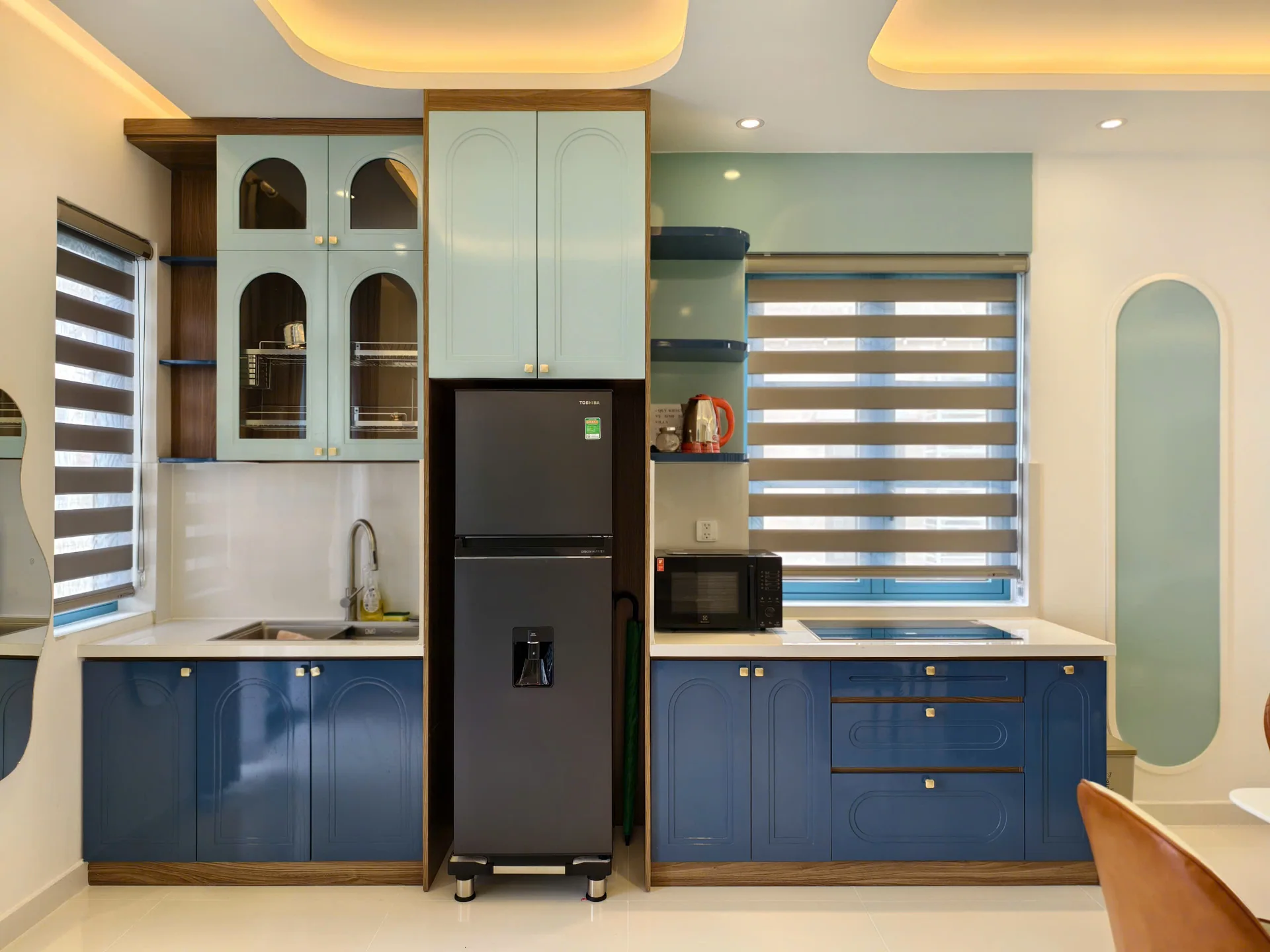 Bếp sau khi làm xong: tủ dưới xanh dương, tủ trên xanh nhạt có ô kính vòm, tủ lạnh đặt giữa