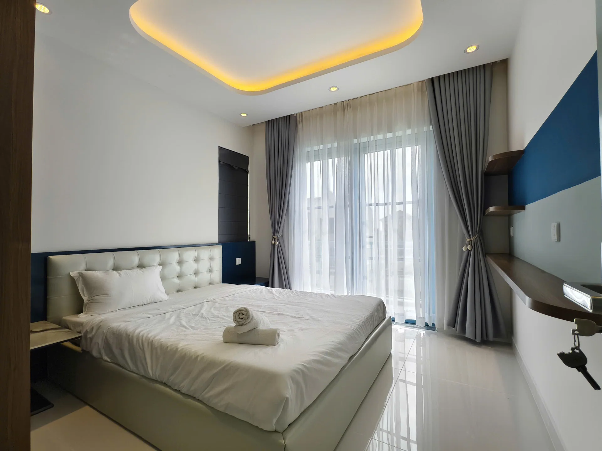 Ảnh thực tế phòng ngủ với giường đôi đầu giường bọc nệm, khăn gấp trên giường, bàn sát tường và cửa kính ra ban công, công trình D.HOUSE tại NovaWorld Phan Thiết