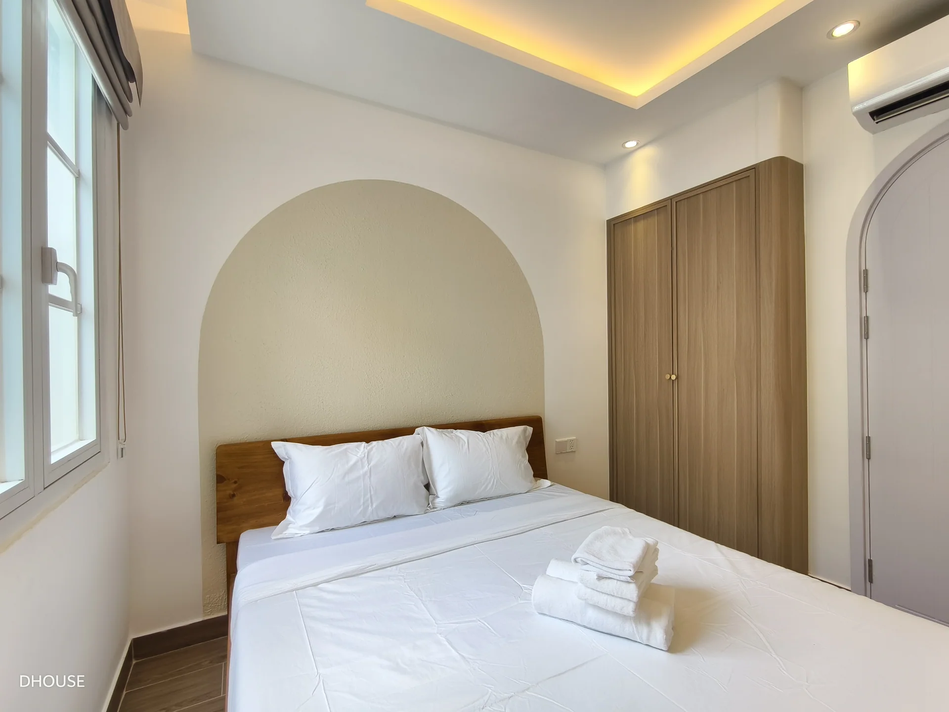 Ảnh thực tế phòng ngủ với giường đặt trong hốc tường vòm, tủ áo gỗ cao sát trần bên phải và đèn hắt trần, công trình D.HOUSE tại NovaWorld Phan Thiết
