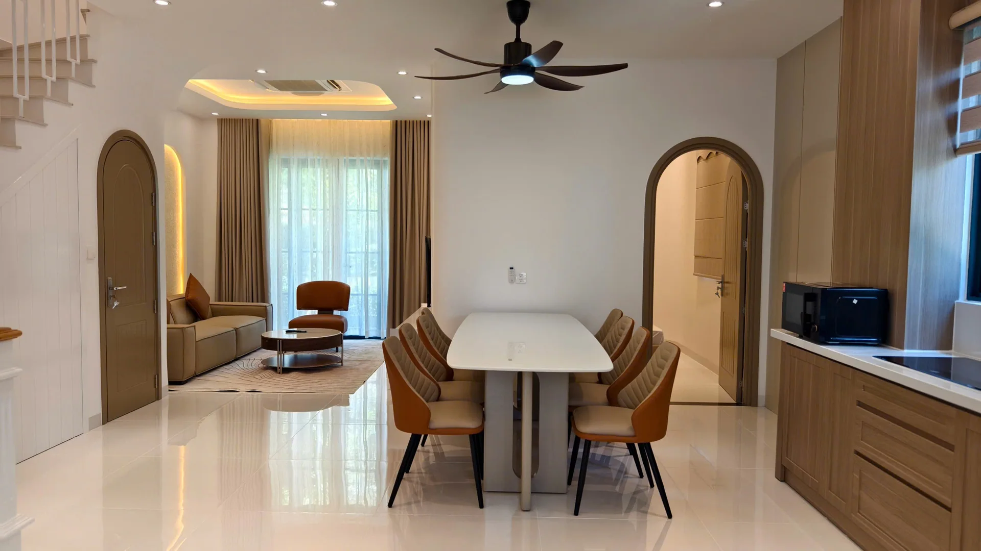Bàn ăn trắng với ghế màu cam, phía sau là sofa phòng khách, cửa vòm và tủ bếp vân gỗ sáng