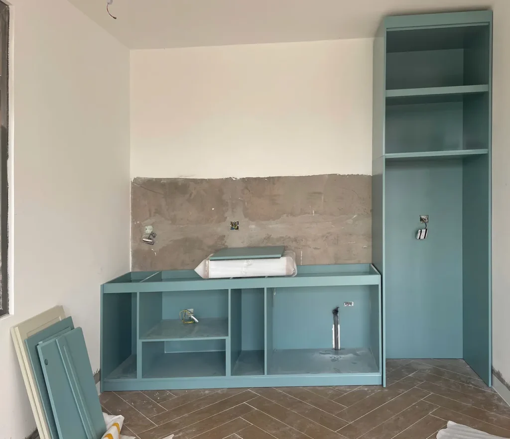 Khung tủ bếp xanh ngọc đã đặt vào vị trí, tường phía sau chưa ốp, cánh tủ còn dựng chờ lắp